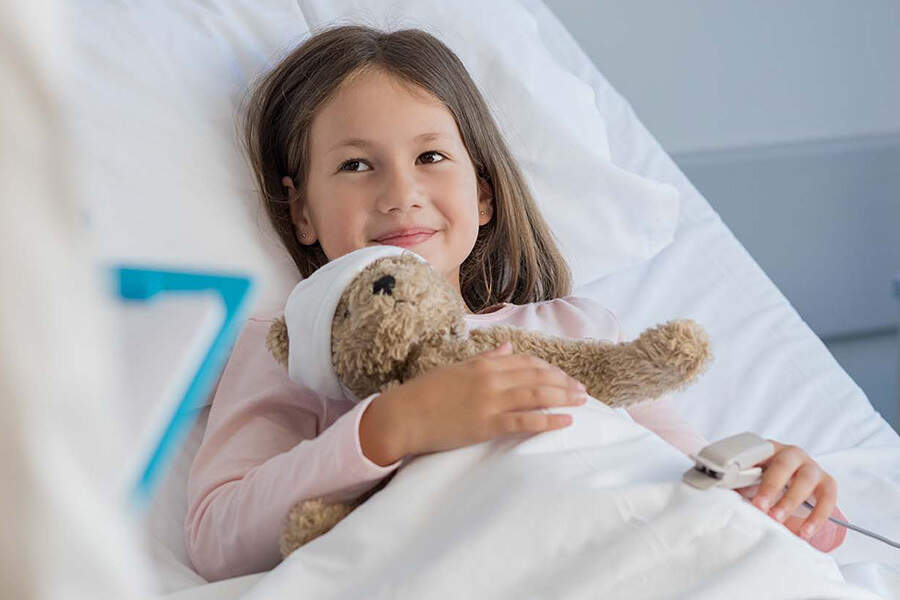 A small girl smiling in a hospital bed while holding a stuffed bear a bandage around his head