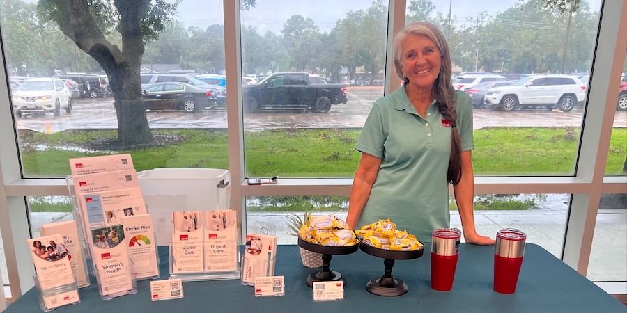 image of a woman standing behind a table that has a teal table cloth. white paper hand outs are displayed on the left side of the table, while wrapped snacks and tumbler cups are on the rigiht side of the table. 