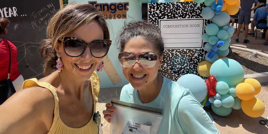 two women posing for a selfie with sunglasses on. they are smiling and standing in front of a backdrop that is themed for back to school with a large composition book and balloons. 