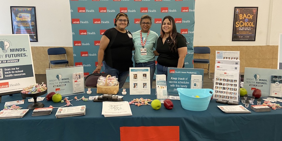 three women standing behind a table filled with back-to-school materials. they are standing in front of a teal backdrop that has the utmb health logo on it. 