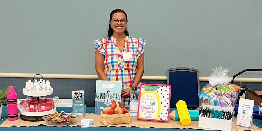 image of table spread at community event with back-to-school-themed materials and giveaways. a hispanic female weearing a white dress with colorful stripes is standing behind the table smiling