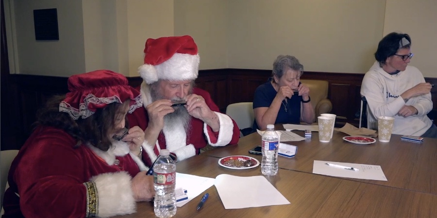 image of four people seated around a table playing the harmonica--two individuals in the foreground are dressed like mr. and mrs. claus the other two are in plain clothes.