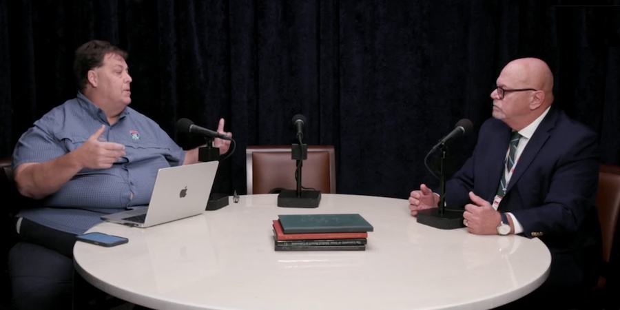 two men sitting at a white round table conversing. they each have microphones in front of them, there's also a third vacant chair with a microphone, too