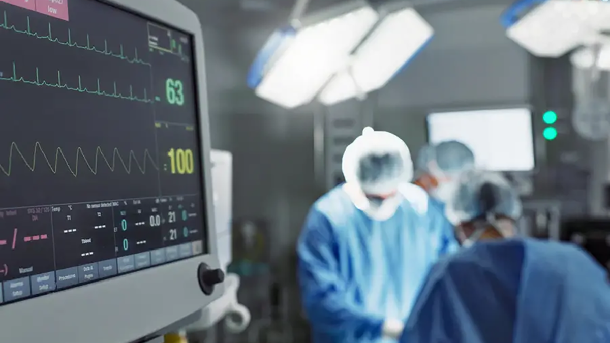 A heart monitor in the foreground with two surgeons performing a procedure in the background