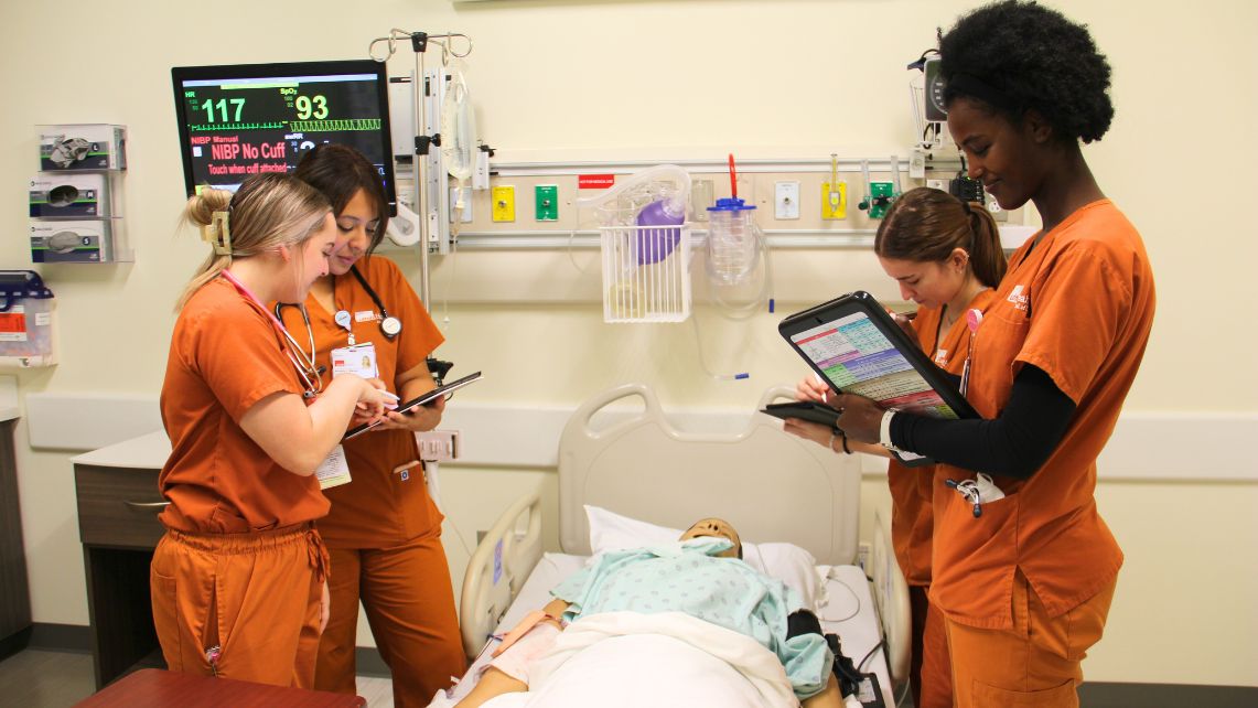 UTMB nursing students in orange scrubs check charts while standing over a mannequin